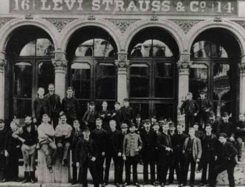 A grainy black and white photo of LS&Co. employees standing in front of a building with four arched windows and a sign reading "Levi Strauss & Co."